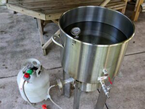 A large stainless-steel pot with water in it attached to a propane tank and a thermometer.